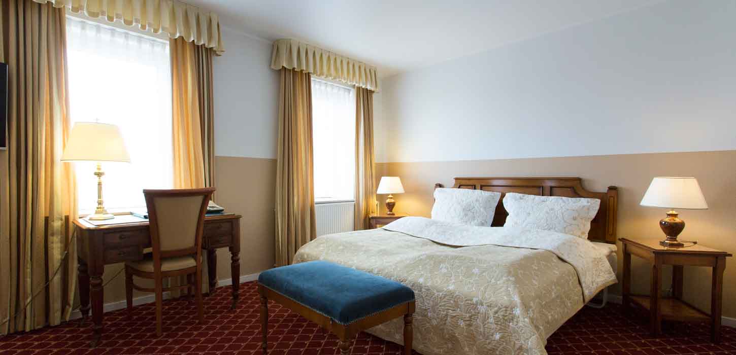 Et hotelværelse med en redt dobbeltseng, tændte lamper på bordene ved siden af sengen og et skrivebord foran et af vinduerne.