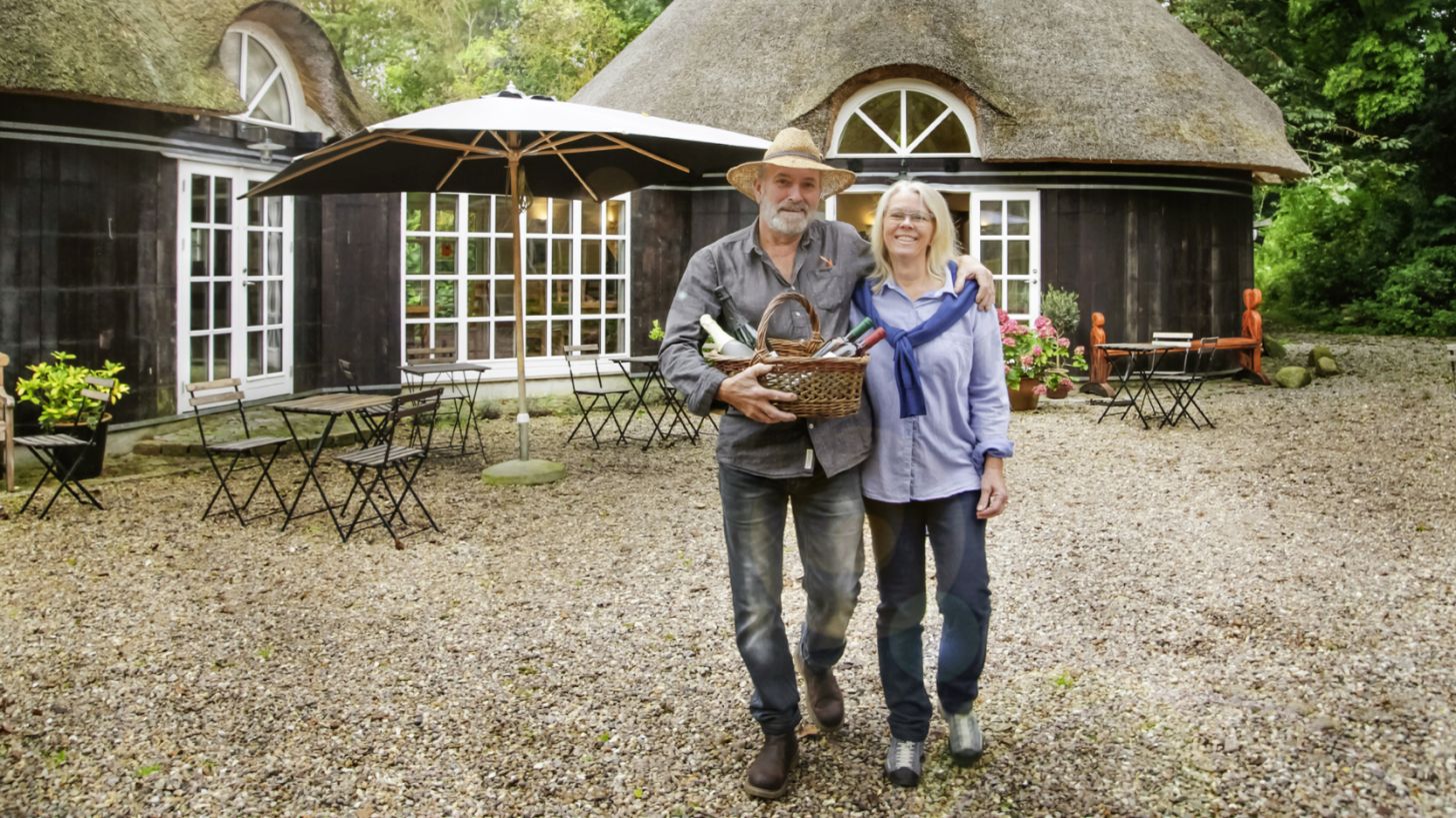 Et ældre par står foran en hyggelig og idyllisk vingård med stråtag.