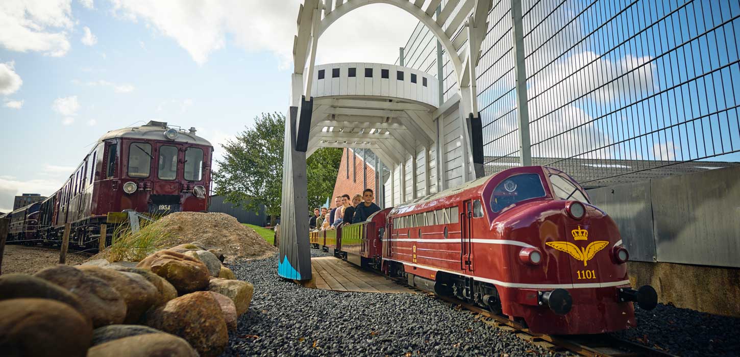 Et tog i normal skala til venstre og til højre ses et lille tog, der trækker børn rundt om en hvid bygning