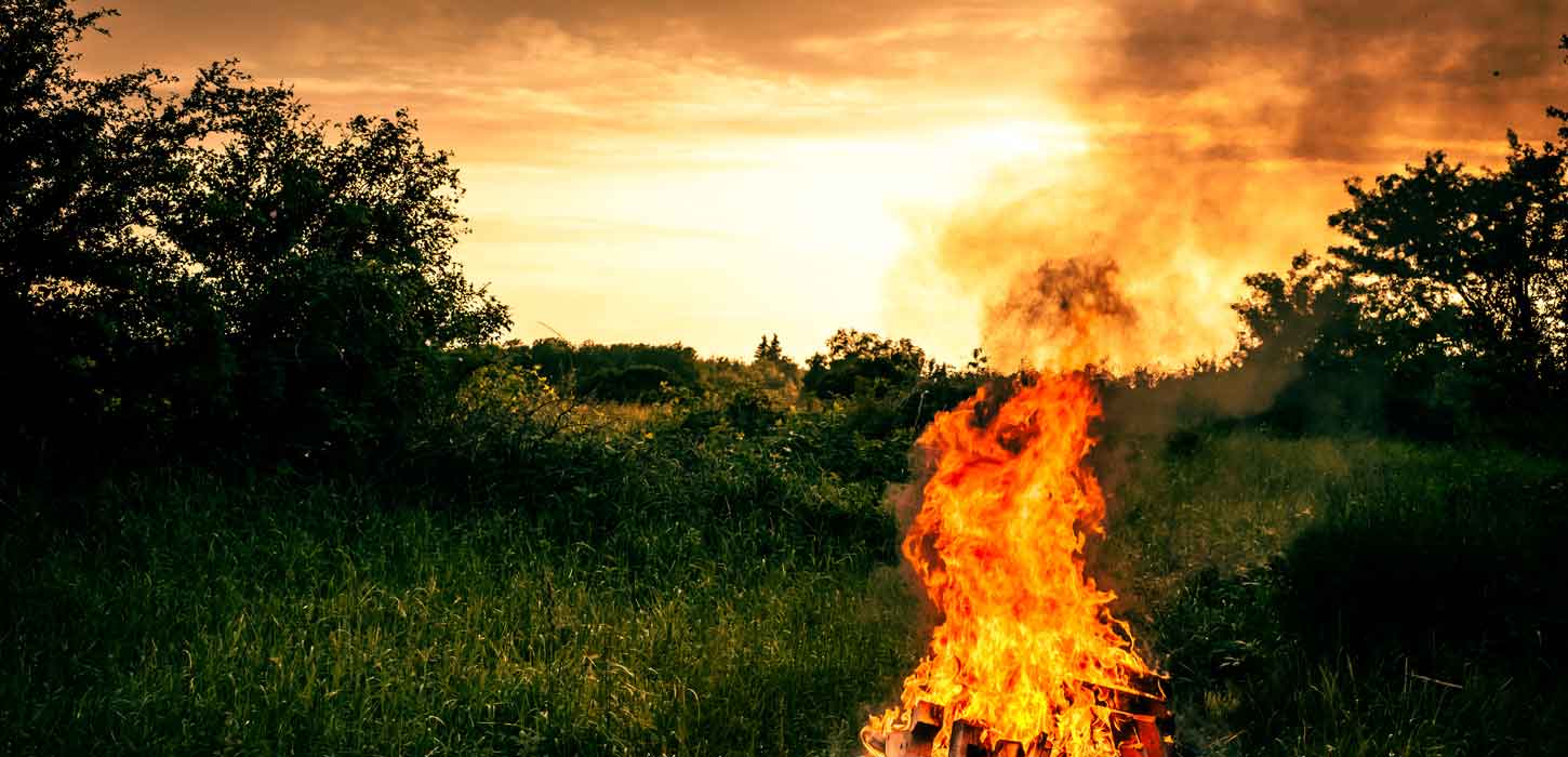 Orange solnedgang og mørkegrønne omgivelser med et stort Sankt Hans-bål i forgrunden.