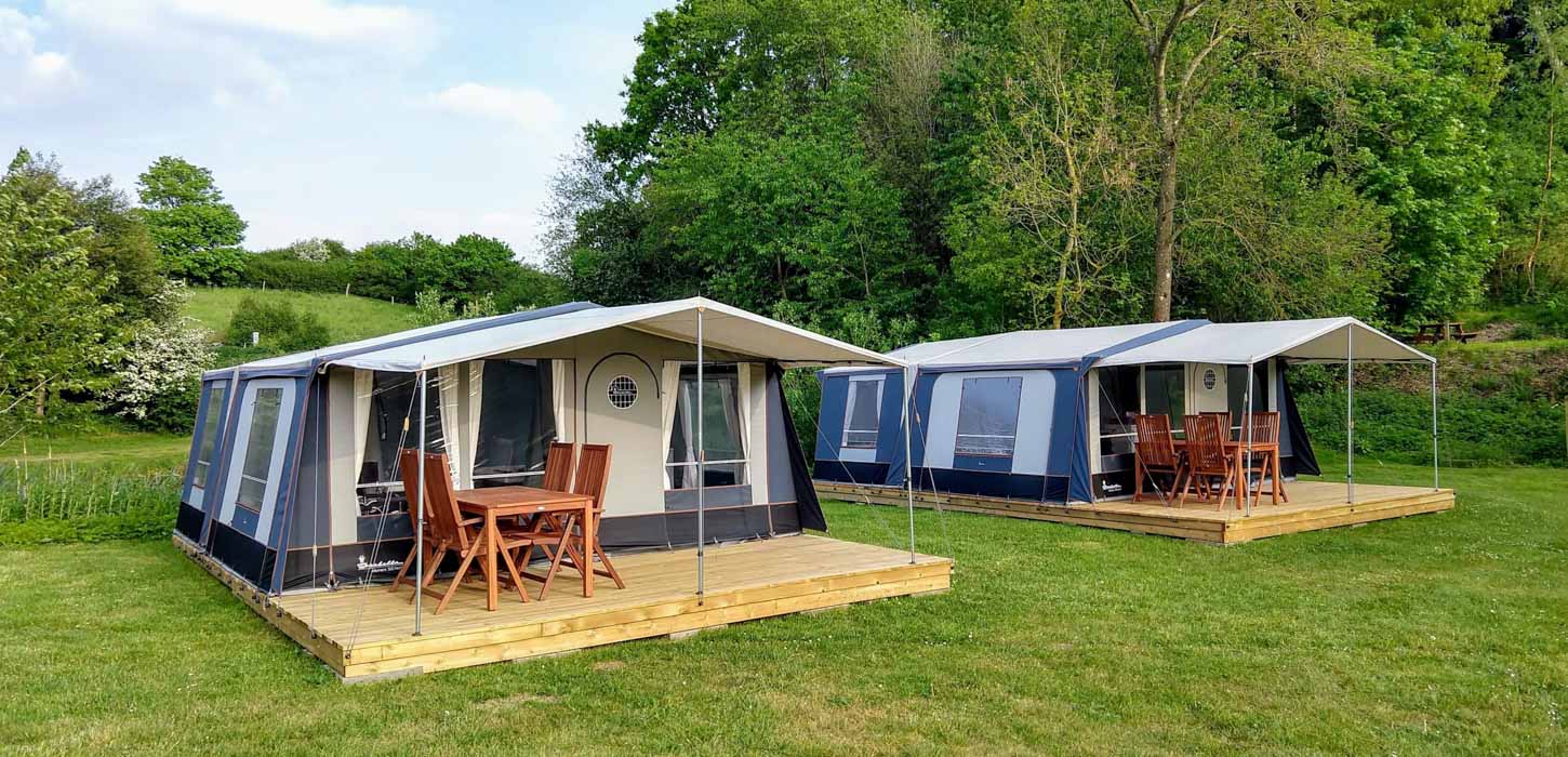 To store campingtelte opstillet på træplatforme på en grøn græsplæne. Bagved ses en bakke med træer og en blå himmel.
