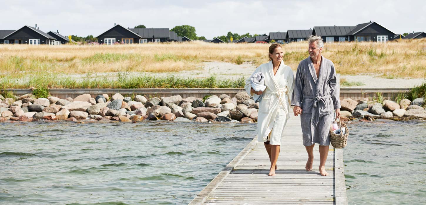 Et par går langs en badebro med sommerhuse i baggrunden og smiler.