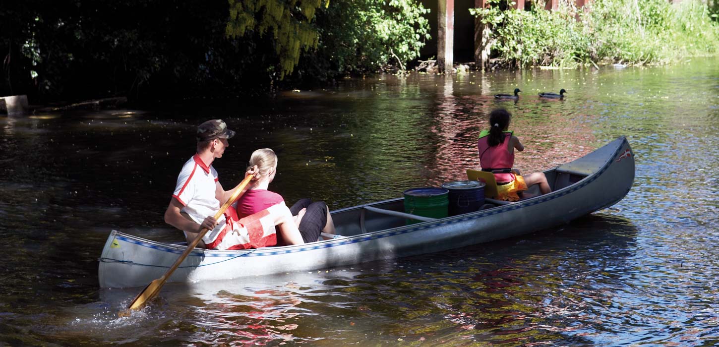 To piger i redningsvest og en mand sejler en grå kano. De sejler på en bred å. I baggrunden ses buske med grønne blade. To ænder svømmer på åen til venstre for kanoen.