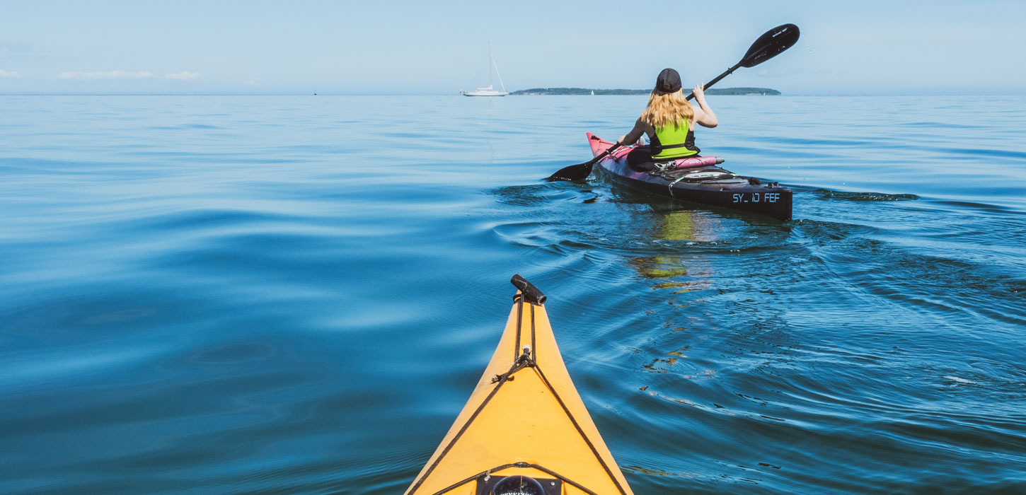Det meste af billedet er dækket af blåt vand. I midten af billedet ses spidsen af en gul kajak. Foran sejler en und lyshåret kvinde en sort kajak. I baggrunden kan man se en sejlbåd og noget land være langt væk.