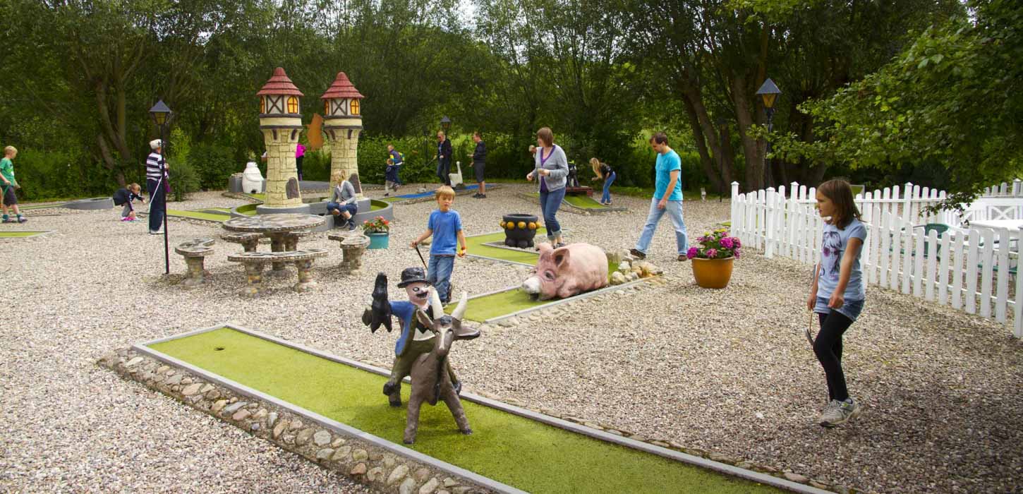 Minigolfbane med folk, der er i gang med at spille. På banen i forgrunden ses en figur af Klodshans på gedebukken.