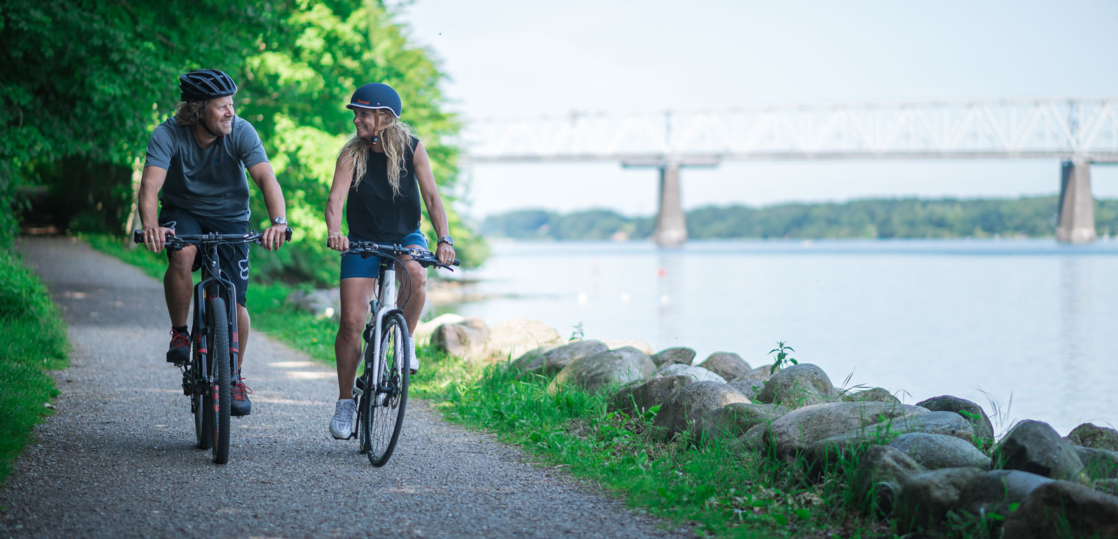 I venstre side cykler et par på hver deres cykel og cykelhjelm. De cykler på en sti der fører ud af en skov. Til højre i billedet adskiller en række af sten havet fra stien. I horisonten kan man se den gamle Lillebæltsbro. 