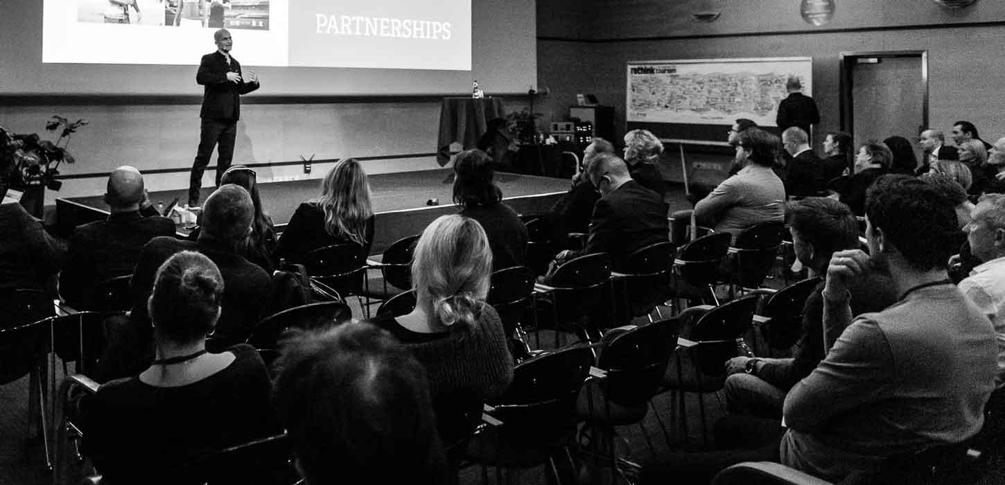 Sort/hvid billede. En folk mennesker sidder på stole og kigger op mod en scene. På scenen står en mand i jakkesæt foran en Powerpoint præsentation og taler.