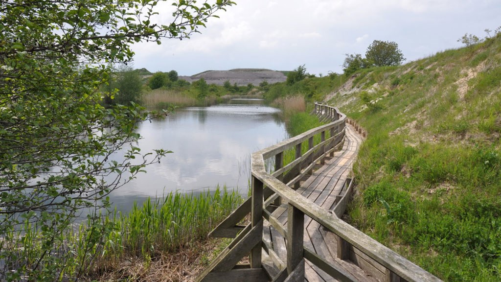 Til venstre løber en lang kalkgrav ud i horisonten. Til højre er en høj fyldt med grøn græs. I mellem dem løber en lang træbro med gelænder i venstre side langs med åen.