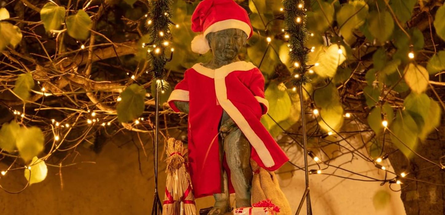 Statuen Mannekin Pis i Bogense klædt i julekostume. I baggrunde ses en lyskæde med lys i og grønne blade.