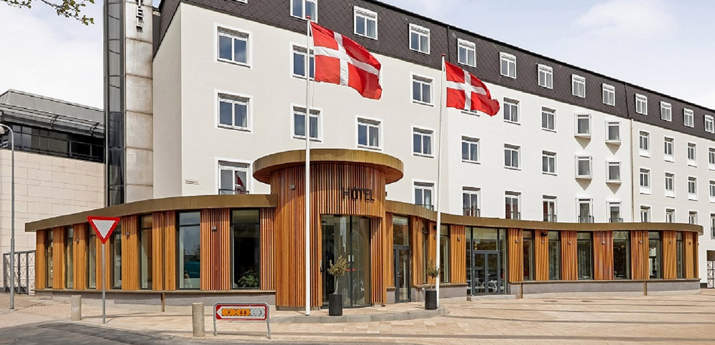 Hvid hotelbygning med sort top. Foran bygningen er et bredt og buet indgangsparti af træ hvor der står Hotel Svendborg. Foran indgangen står to flagstange med det danske flag.