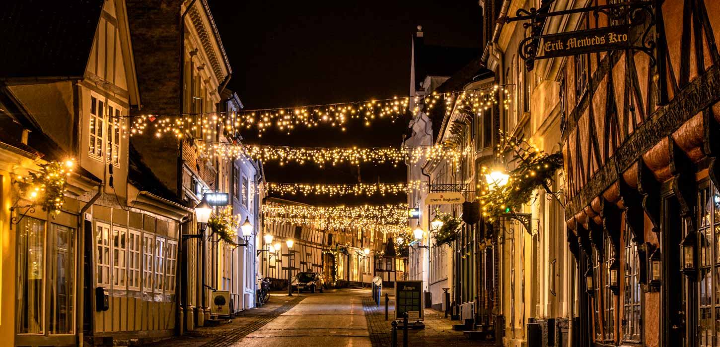 Et billede af en brostensgade i Bogense. På hver side af gaden er der forskellig farvede bindingsværkshuse. Mellem husene hænger julelys og lyser gaden op. 