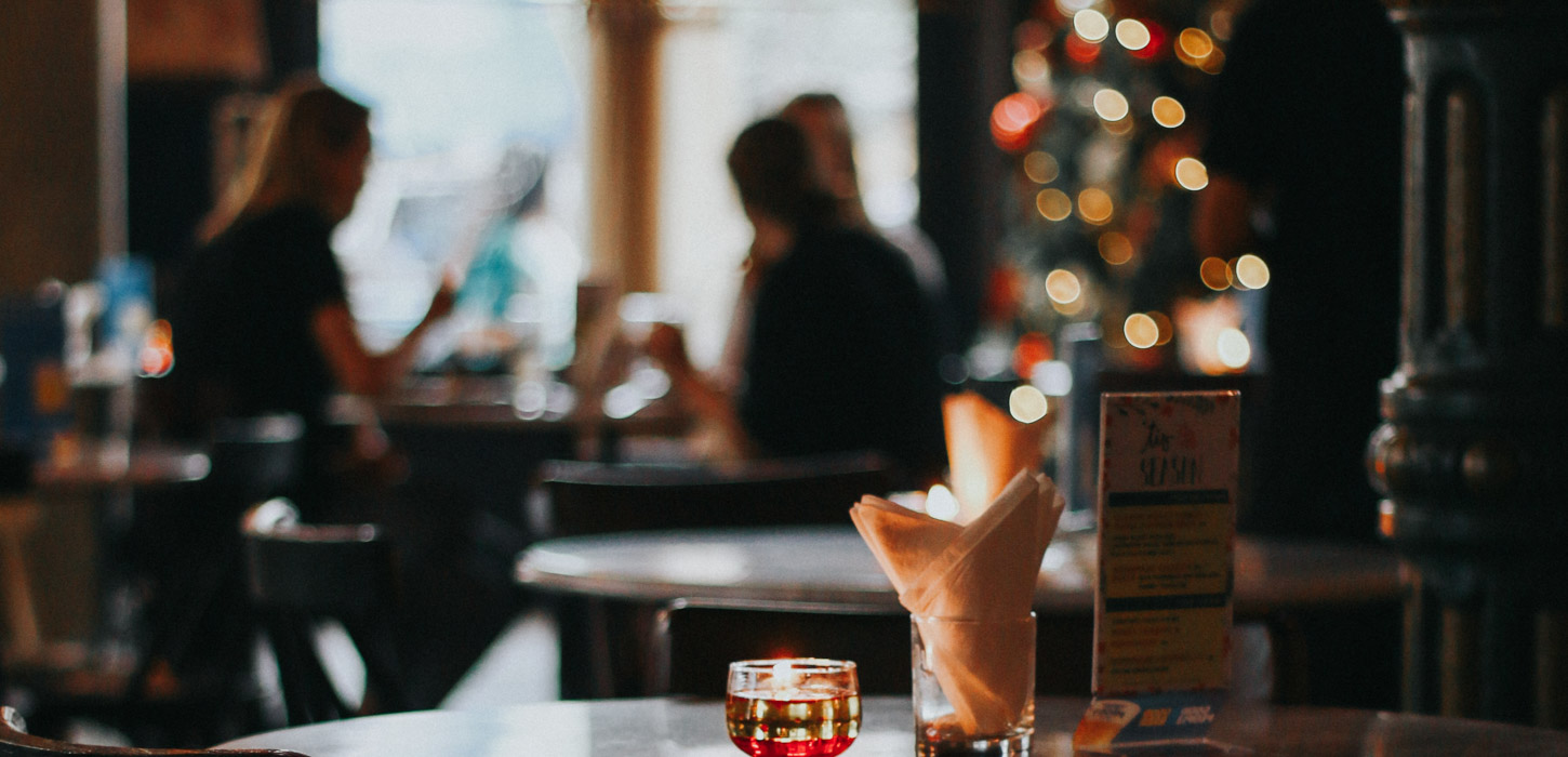 I forgrunden er der fokus på et lille fyrfadslys, et glas med en foldet serviet og et menukort. I baggrunden står der to runde borde med stole omkring. Ved det ene bord sidder tre slørede mennesker og læser menukort. Ved siden af dem står et pyntet juletræ og lyser.