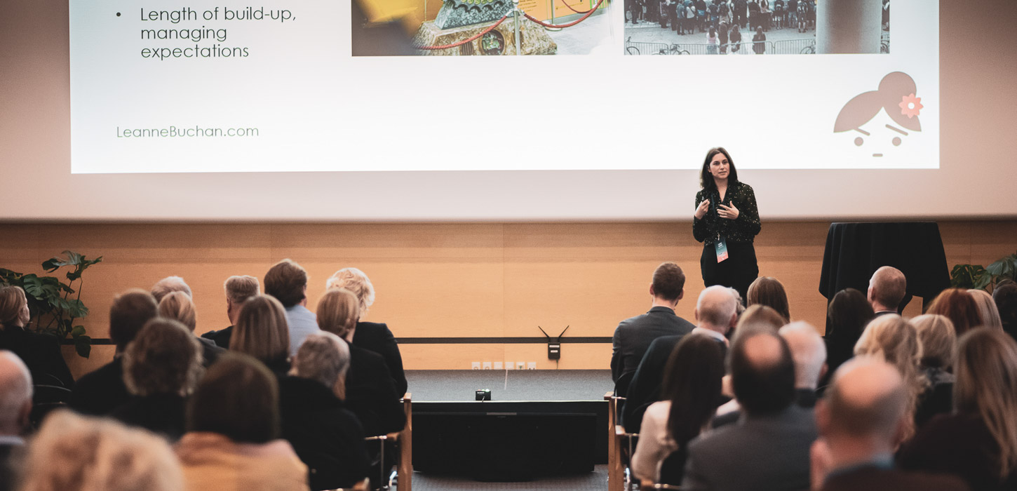 En masse mennesker sidder og kigger op på en kvindelig foredragsholder, som står og snakker.  Bag hende er en stor skærm med et powerpointslide.