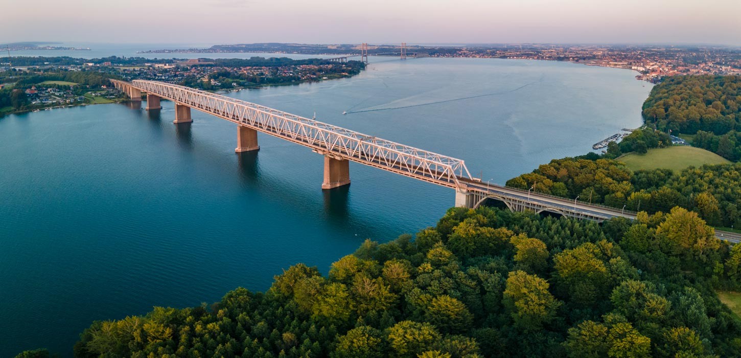Et dronebillede af Lillebæltsbroen der strækker sig over det blå hav. På hver side ses store mængder af træer og små bygninger set oppefra.