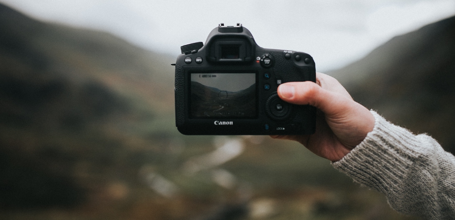 Et nærbillede af en hånd med grå sweater der holder et spejlreflekskamera fra Canon. Baggrunden er sløret men viser et grønt bakket landskab.