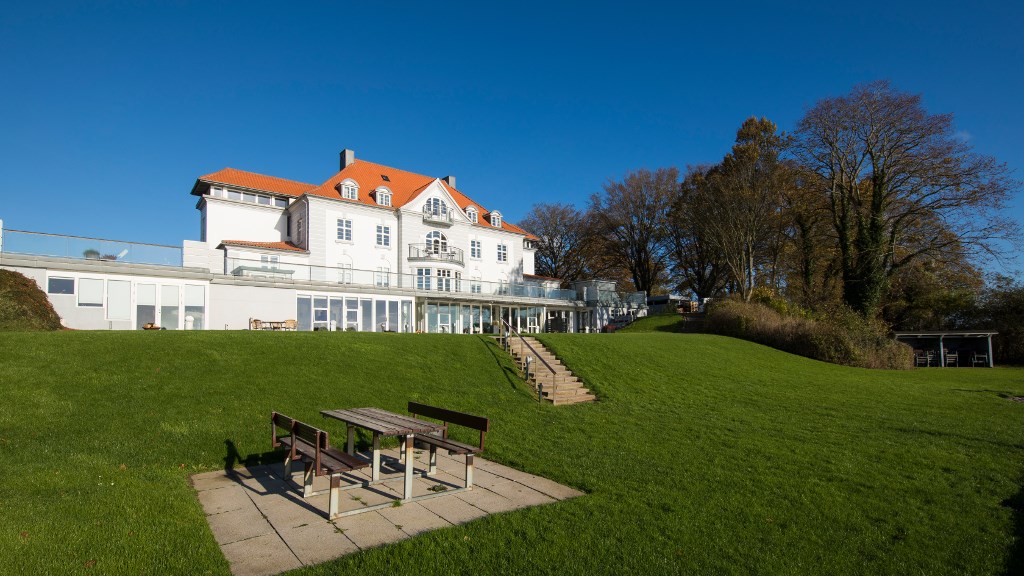 Stort, hvidt hotel med rødt tag beliggende i en stor have. Bænkesæt i forgrunden.