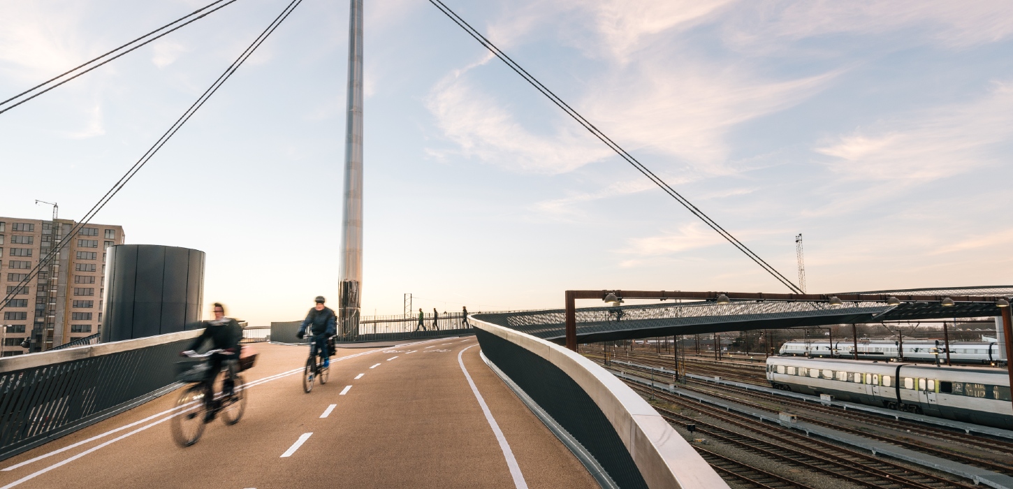 Byens Bro i Odense med to cyklister i fart på vej henover broen. Etagebygning i baggrunden, og togene nede på skinnerne til højre i billedet.