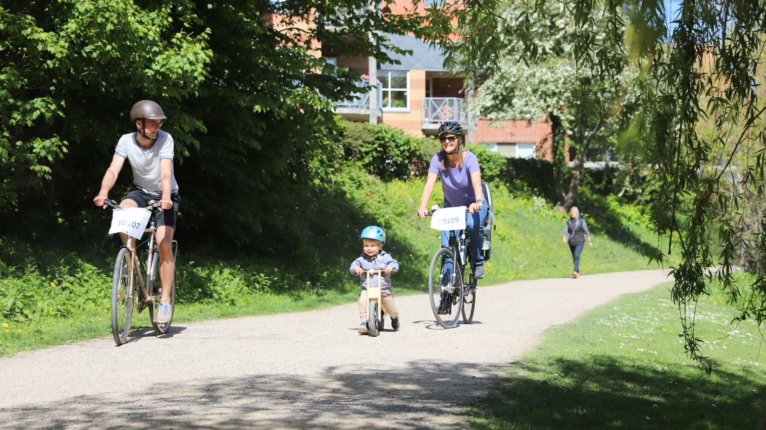 En mand, en kvinde og et lille barn på løbecykel cykler hen ad en solbeskinnet sti i grønne omgivelser.