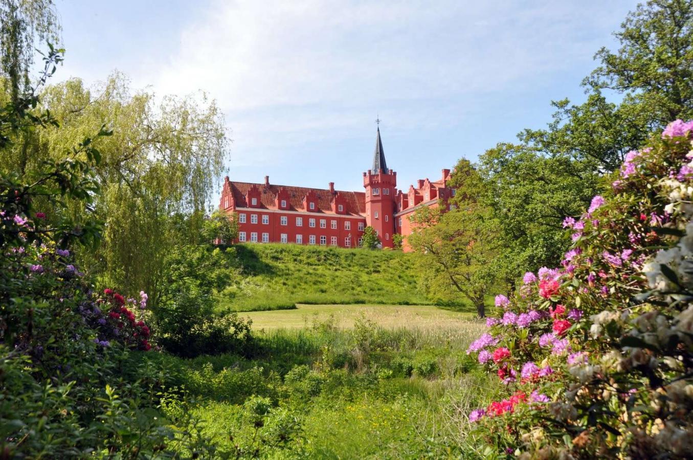 Rødt slot ses på afstand med flot spir og omringet af grønne buske og blomster