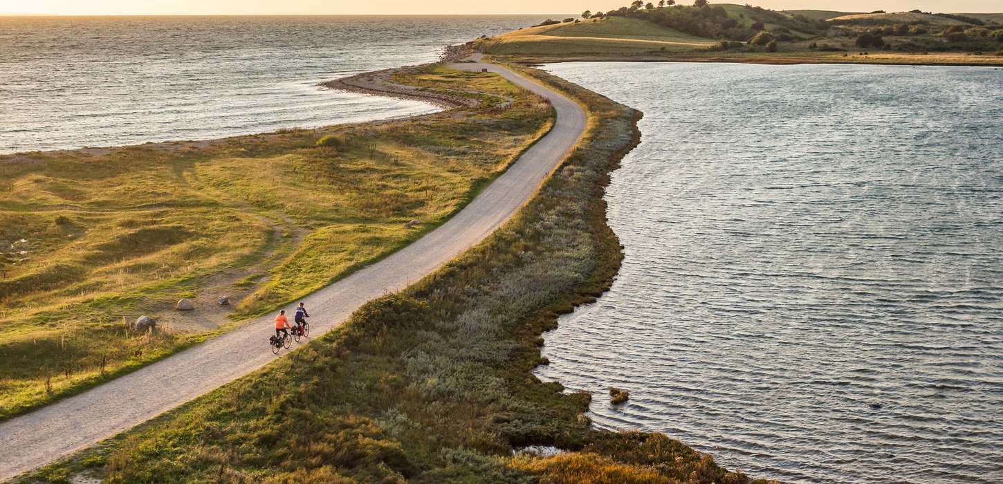 Luftfoto af to cyklister på smal vej ud af tange med vand på begge sider og land i baggrunden.