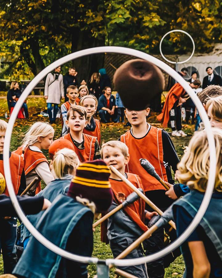 Gennem en hvid hullahopring kan man se en flok børn. Den ene halvdel har røde veste på og den anden blå. De har alle sammen kosteskafter mellem benene og kaster en bold. I baggrunden er forældre tilskuer, mens de står under et kæmpe træ. Børnene spille spillet quidditch fra Harry Potter.