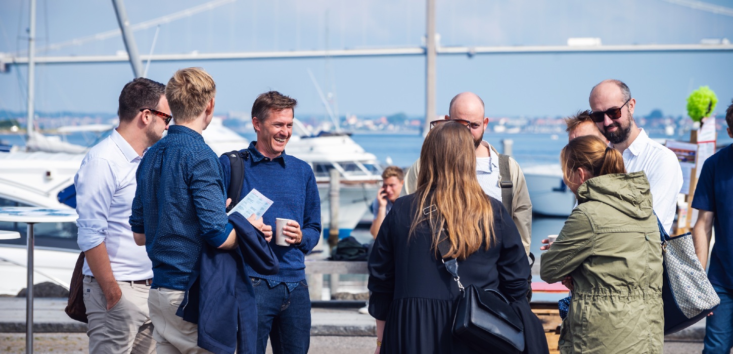 I forgrunden står otte mennesker i en cirkel og snakker sammen. I baggrunden ses en masse skibe der ligger i havn. I horisonten kan man se Lillebælt broen.