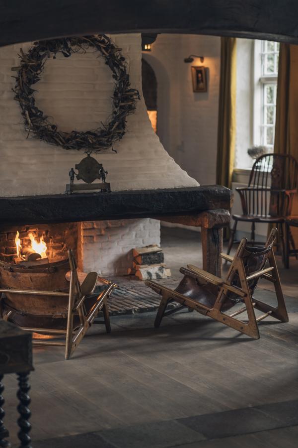 To tomme stole foran åben kamin med ild i et rum med hvide murstensvægge. Lyset står ind ad et vindue i højre side.