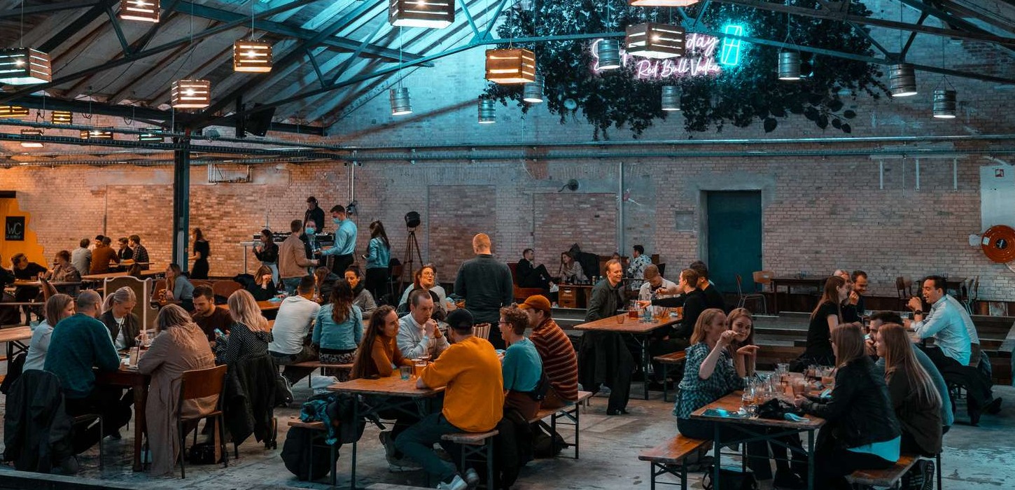 En masse mennesker sidder ved borde i stort lokale med rå murstensvægge og lamper i loftet.