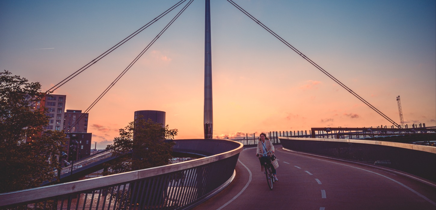 Byens bro i Odense vist i en flot solnedgang med gule og orange nuancer. En cyklist kommer cyklende på vej ned af broen.