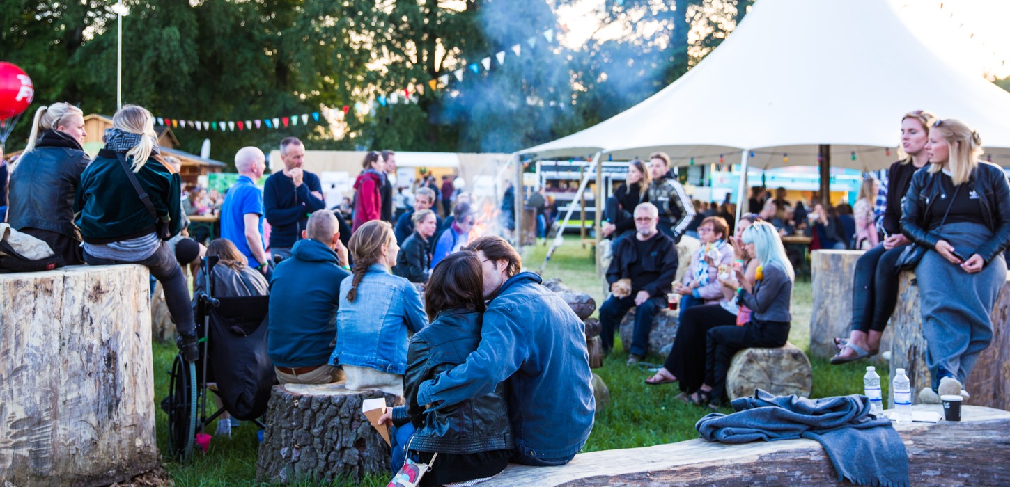 Foran et telt og træer sidder en stor gruppe mennesker på træstubbe rundt om et bål.