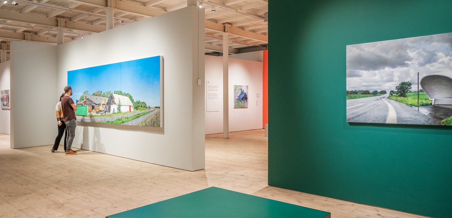 Udstillingslokale med en hvid vær og en grøn væg med store malerier på begge. To personer står og betragter billedet på den hvide væg.