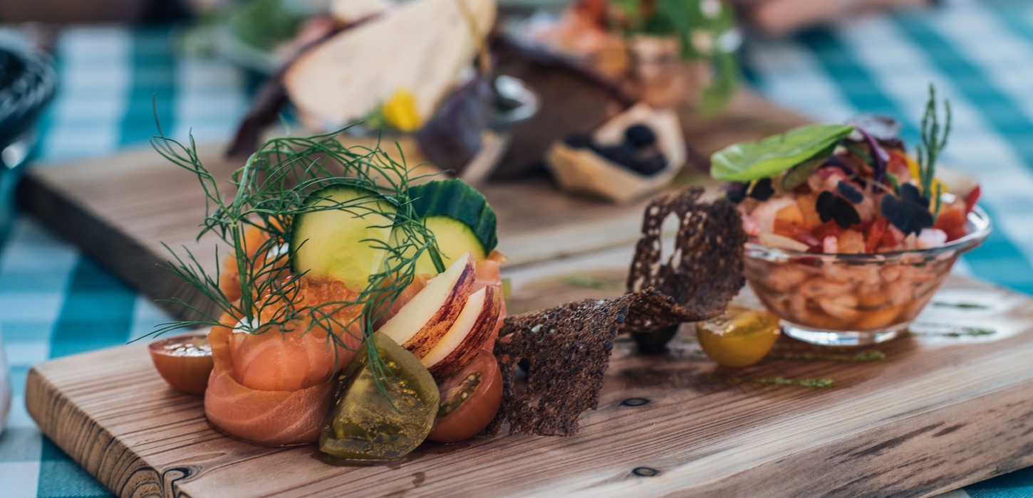 På en træskærebræt. Til venstre på brættet ses en blomst lavet af laks pyntet med agurk, dild og tomater. Til højre ses en glasskål fyldt med rejer og pyntet med grønt. Imellem de to retter ligger en buet rygbrødschips. I baggrunden kan et sløret bræt ses. De står begge på en hvid og blåternet dug.