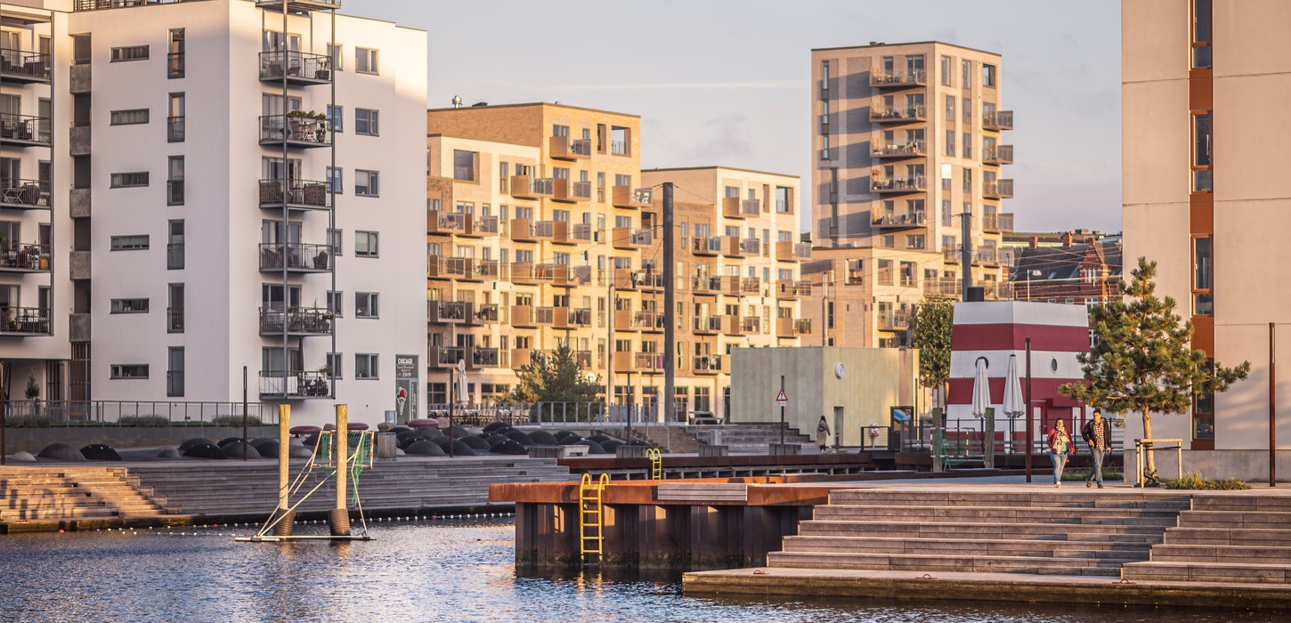 Sollys skinner på lejlighederne ved Odense havn. To mennesker sig en tur ved havnen i det flotte vejr.