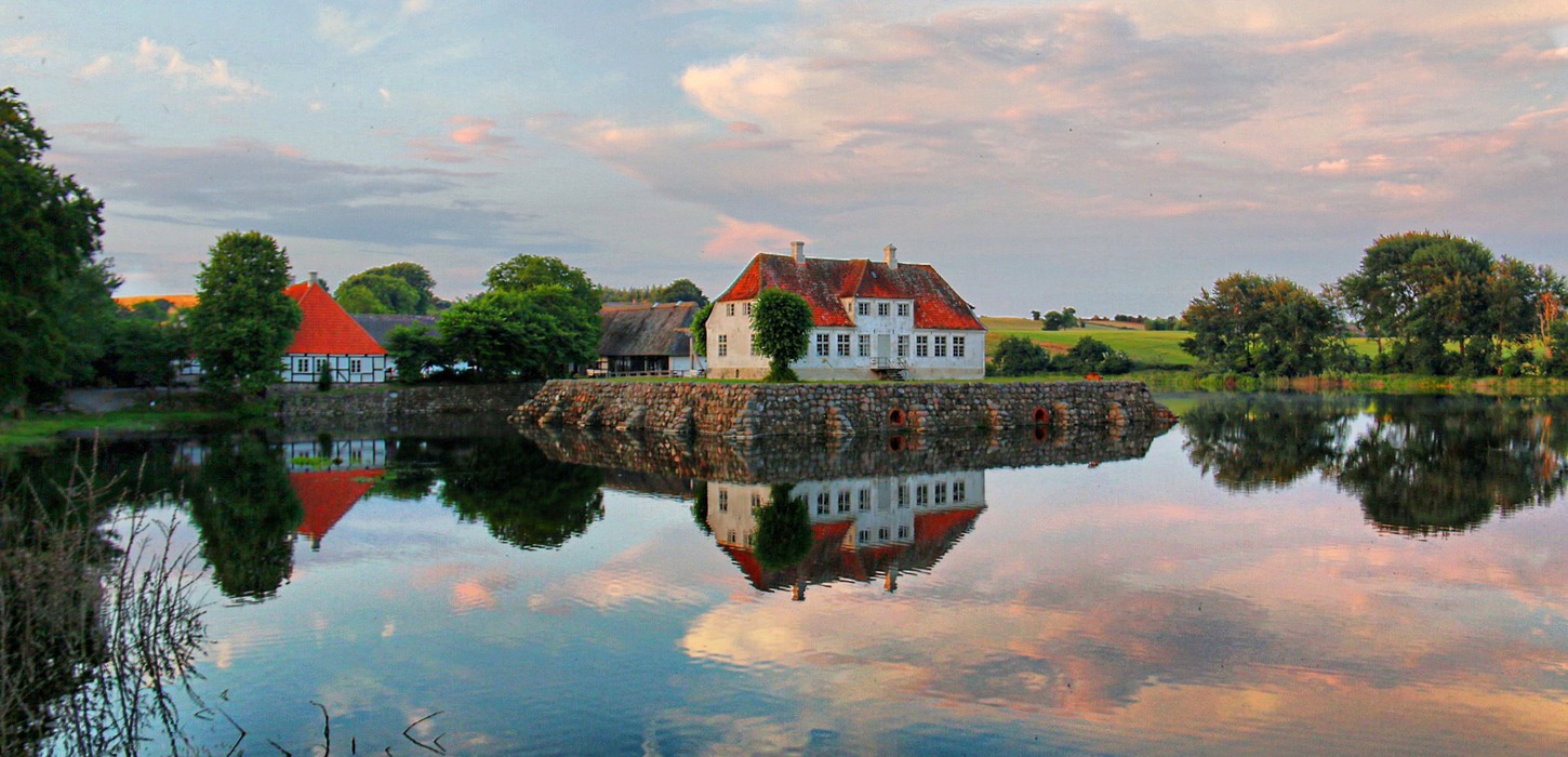Flot blankt spejlbillede af en hvid gård med rødt tag i grønne omgivelser og lyserøde skyer.