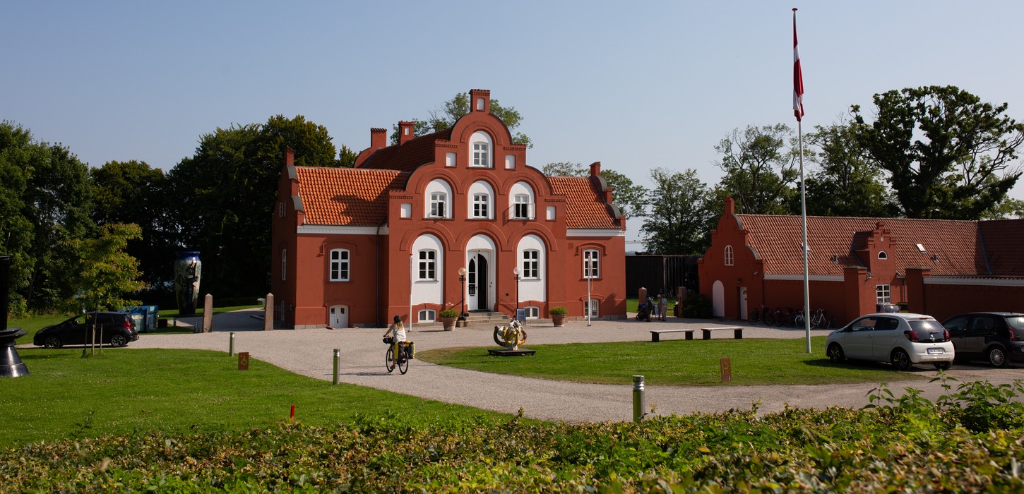 Prægtig rød bygning med gårdsplads. Flaget er hejst foran bygningen og der holder et par parkerede biler.