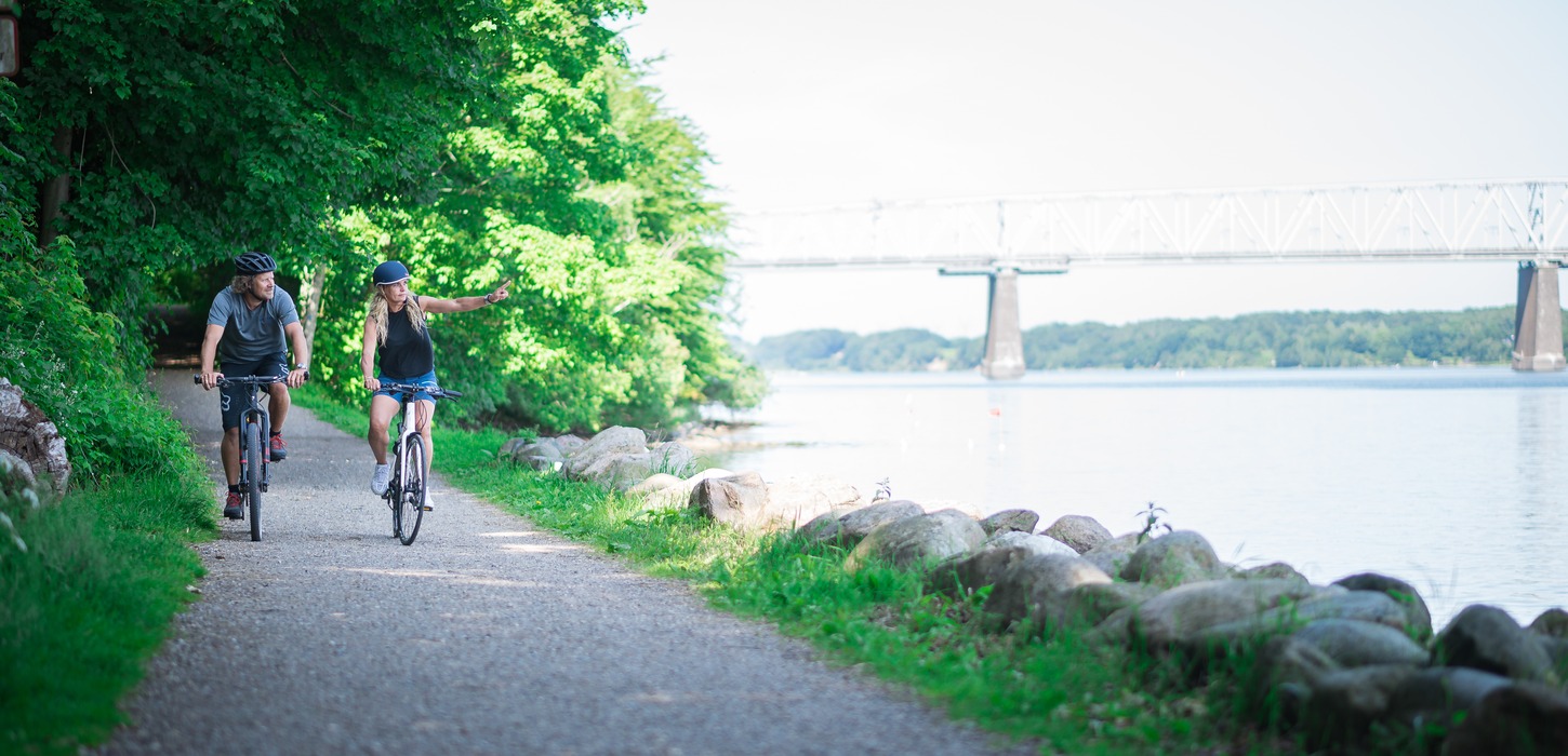To cyklister kommer kørende ad sti langs med vandet. Den ene peger ud over vandet. En bro anes i baggrunden.
