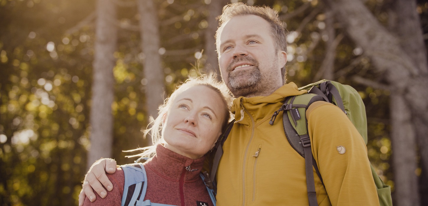 Closeup af kvinde og mand, der holder om hinanden med skov i baggrunden og sol i ansigterne.