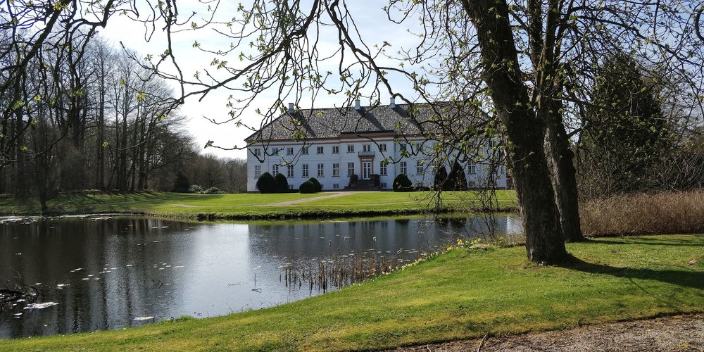 I baggrunden af en blank sø, et par træer og en grøn græsplæne ses Krengerup gods. Godset er hvidt med sort tag, og foran står et par buske.