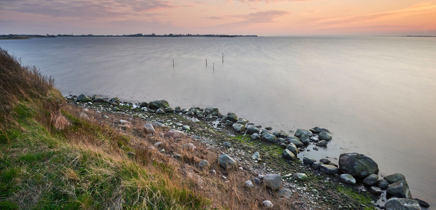 Et billede af hav med en himmel i horisonten der er oplyst i en solnedgangs farver. I forgrunden ses kyst med sten der adskiller græs fra havet.