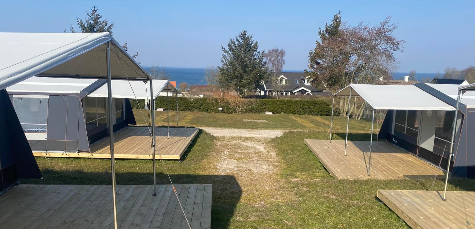 En række ens udseende campingvogne med fortelte og træterrasser med træer, hæk og vand i baggrunden.