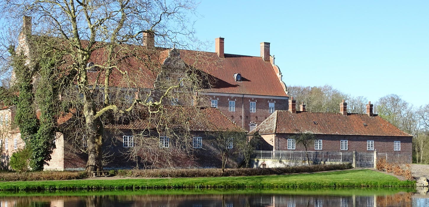 Prægtige rød herregård med røde tage. Foran bygningerne er der vand, grønt græs og et stort træ med krogede grene .