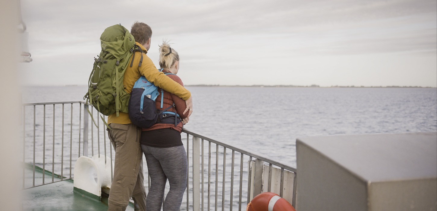 Et par i vandretøj og med rygsække står sammen og kigger ud på havet. De står op af et rækkeværk på bagdækket af en færge.