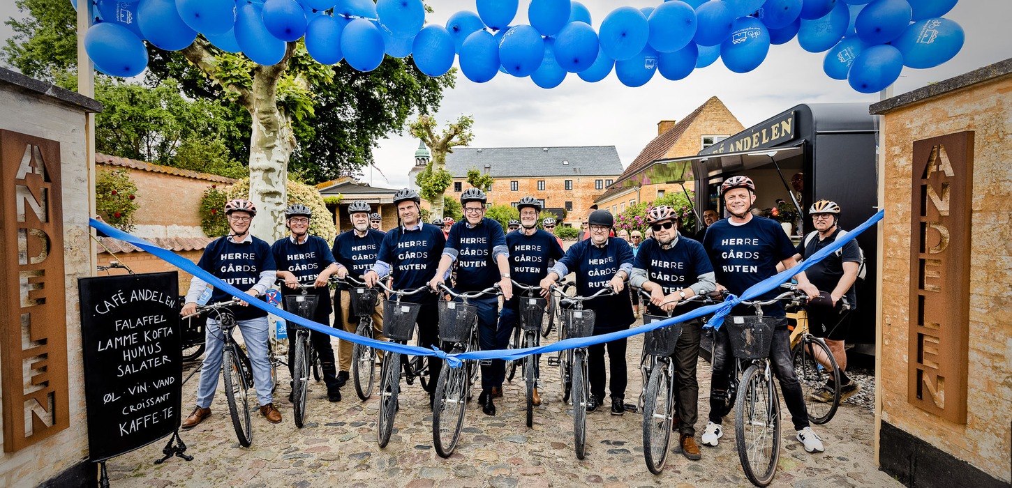 De fynske borgmestre stå klar på cyklen på ræd og række foran et blår bånd ved Andelen på Ærø. En port af blå balloner ses henover dem.
