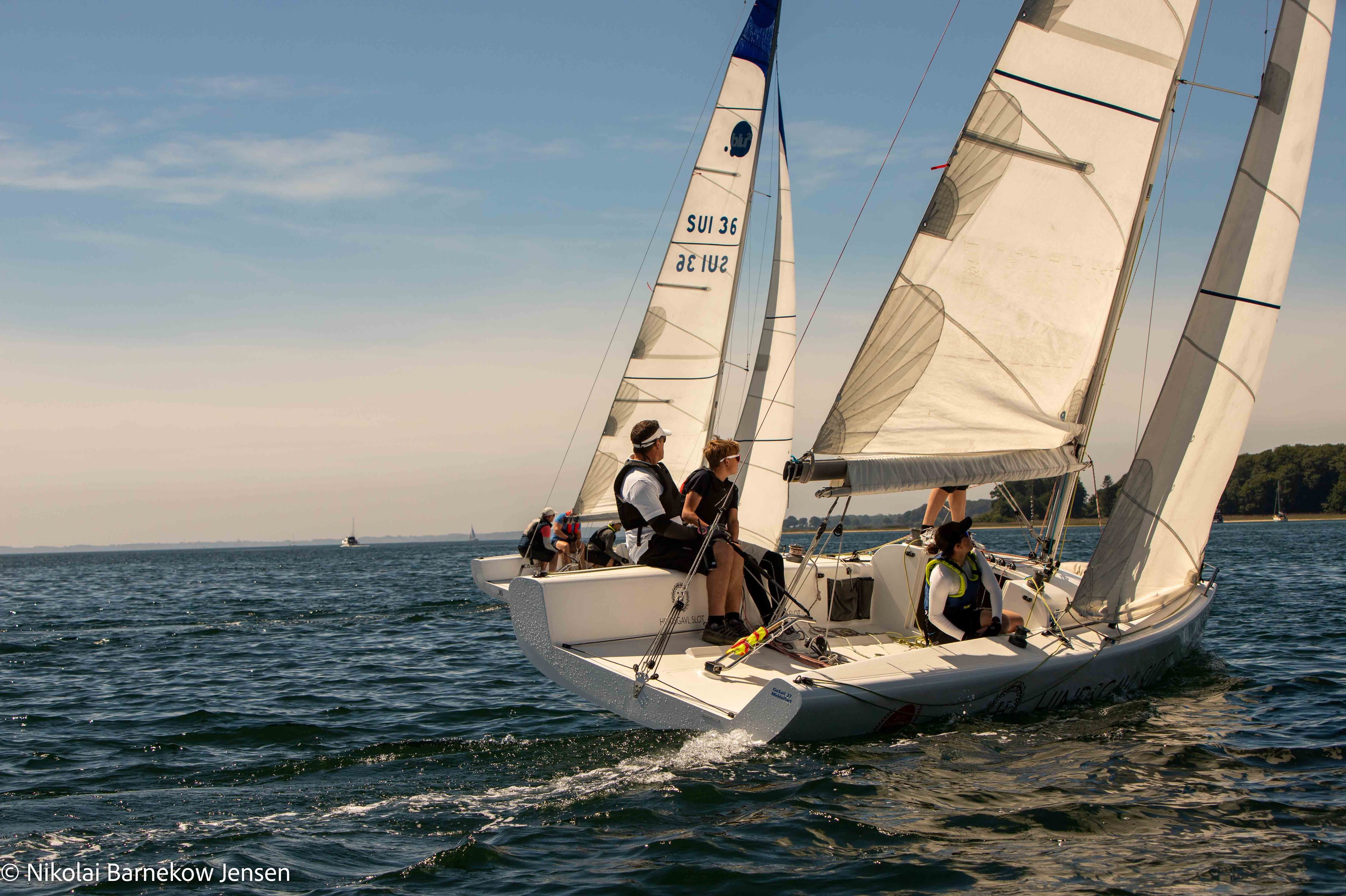 På åbent hav sejler en hvid konkurrence sejlbåd. De har fire sejl slået op, og fire mennesker hjælper alle med at sejle båden. I horisonten glider himmel og hav sammen.