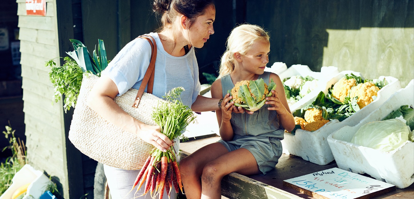 Kvinde og pige ved en åben vejbod med grøntsager. Piger holder et blomkål i hånden, kvinden et bundt gulerødder.