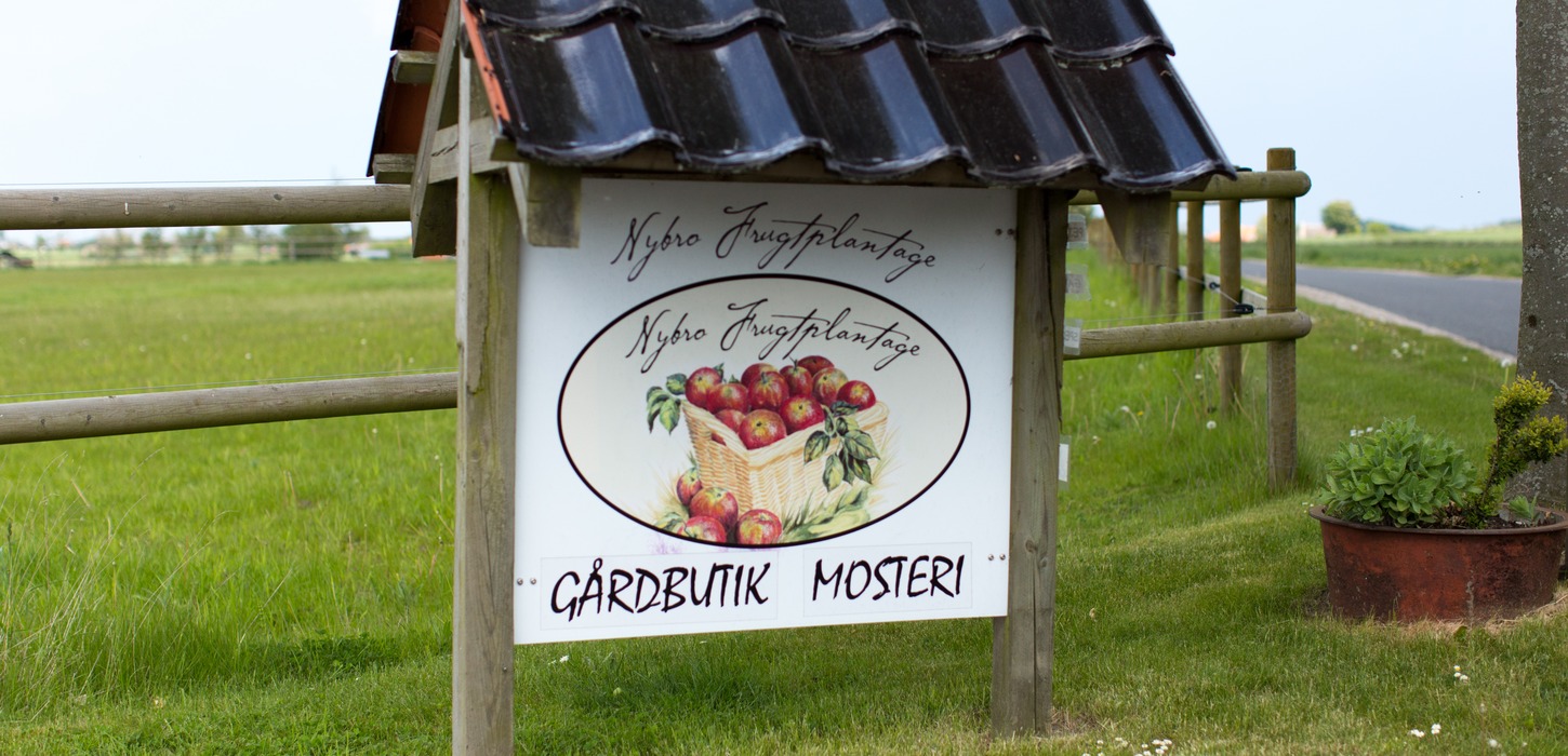 Skilt i vejkanten med et billede af æbler og teksten "Nybro Frugtplantage - Gårdbutik Mosteri"
