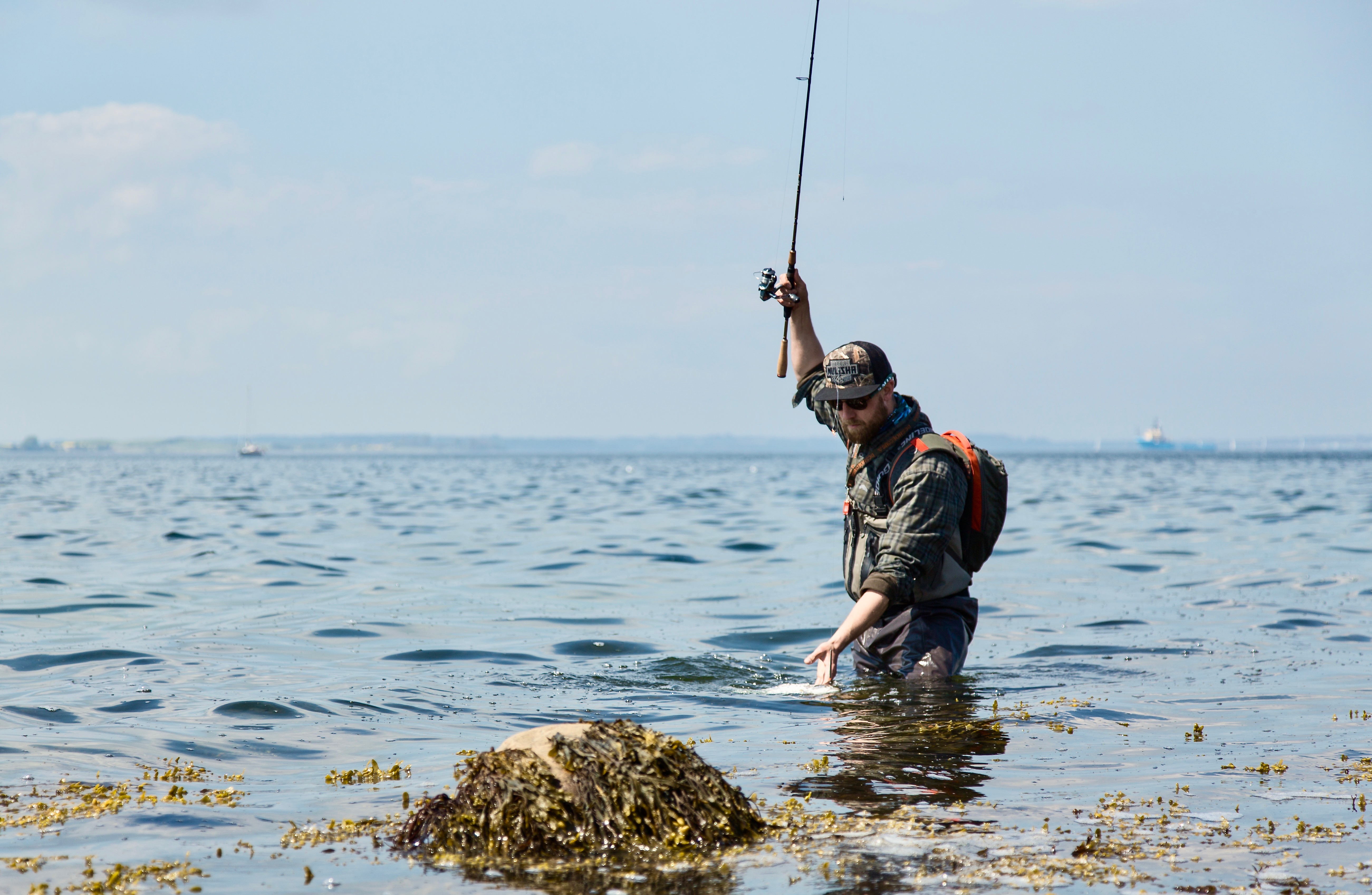 En fisker står med vand til lårene. Han har fiskestangen løftet lige op i luften og er ved at tage en fisk op af vandet.
