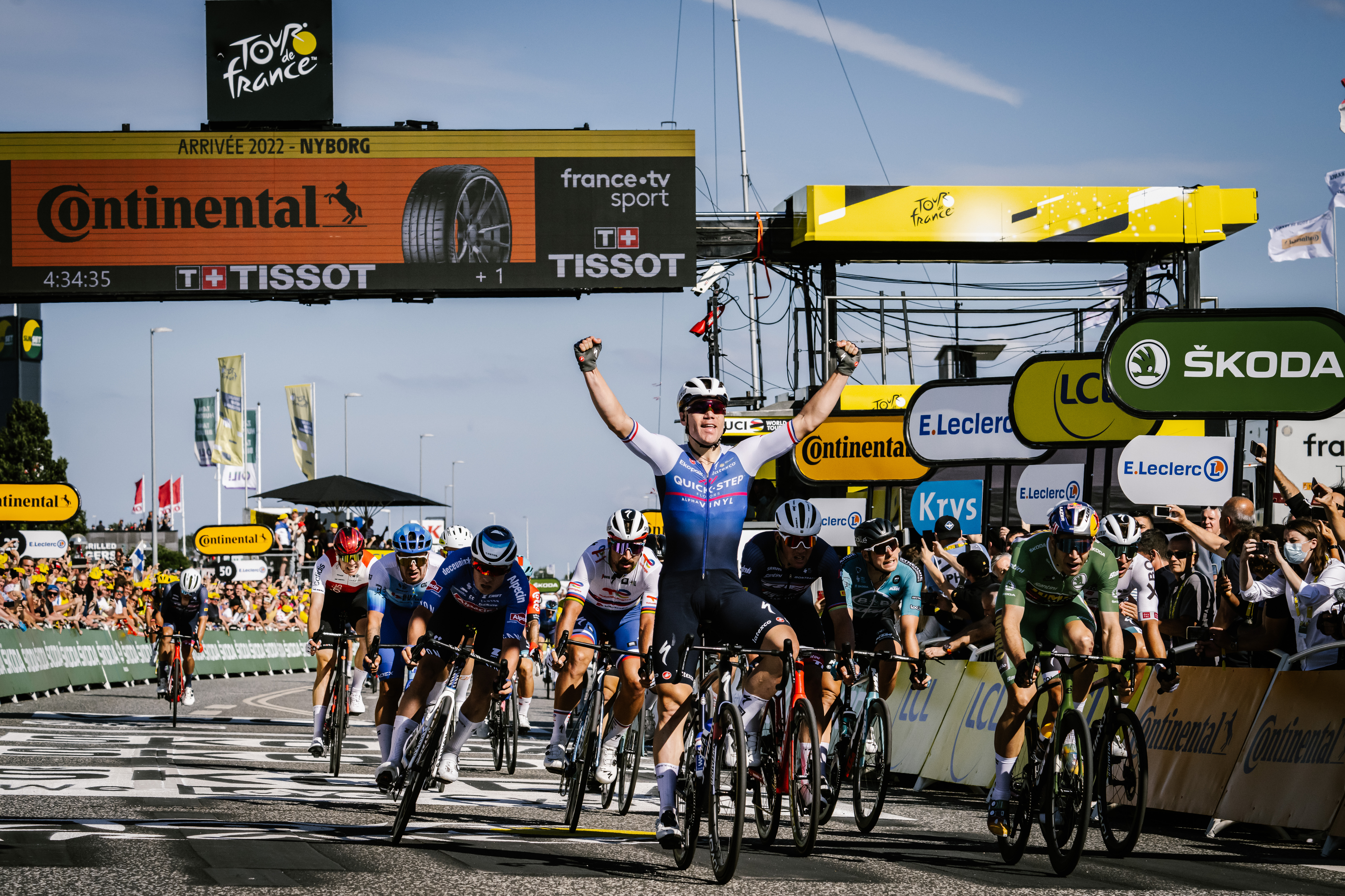 En masse cykelryttere til Tour de France fotograferet ved målområdet. En rytter har armene oppe i sejr.