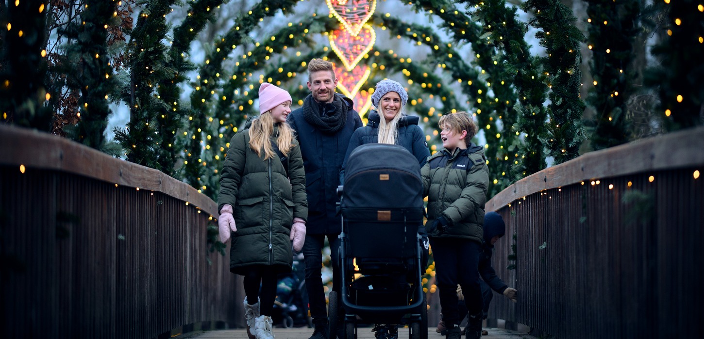 På en træbro står en familie med tre brøn i vintertøj. Foran moren står der en barnevogn. Over hele broen er der buer med gran, lyskæder og i midten et lysende julehjerte.