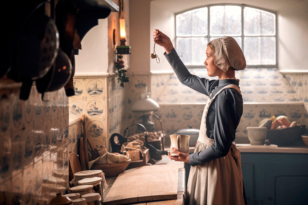 Et hjørne af et gammeldags køkken. Køkkenskabene er blå, bordpladen er af lyst træ, og i den nederste del af væggen er der lysebrund klinker med blåt mønstre. På bordpladen står en kurv med grøntsager, og en masse små glas med mad. Ved køkkenbordet står en und kvinde i stuepigetøj og kigger på en halskæde med hjertevedhæng.